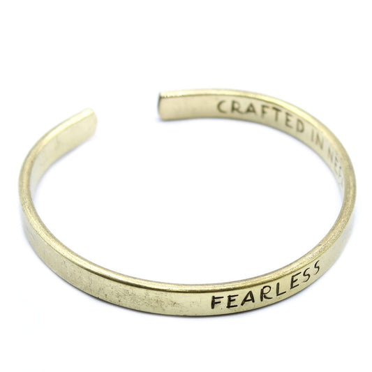 A slightly open, golden-colored metal bracelet displays the text "CRAFTED IN NE... FEARLESS" along its inner surface against a plain white background.