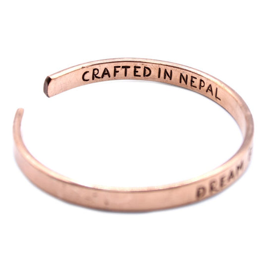 A rose gold metal bracelet curves in a C-shape against a white background, displaying engraved text that reads: "CRAFTED IN NEPAL" and "DREAM."