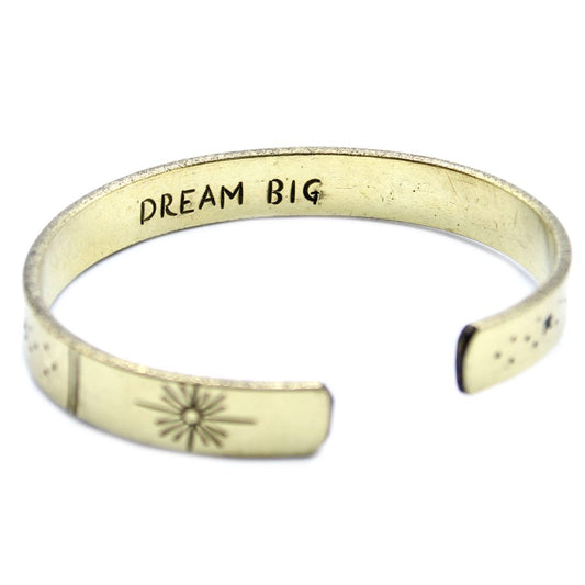A gold-toned metal cuff, engraved with "DREAM BIG" on the top and sun/star designs on each end, rests on a white surface.