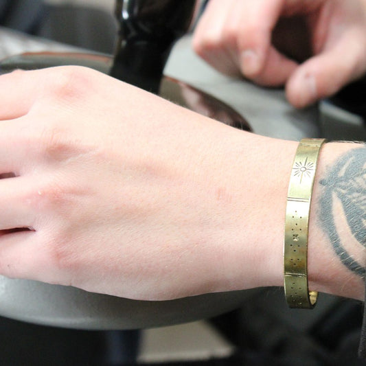 A wrist wearing a gold-colored bracelet with star engravings rests near a blurred, dark object. The arm has a visible tattoo. Another hand is blurred in the background.