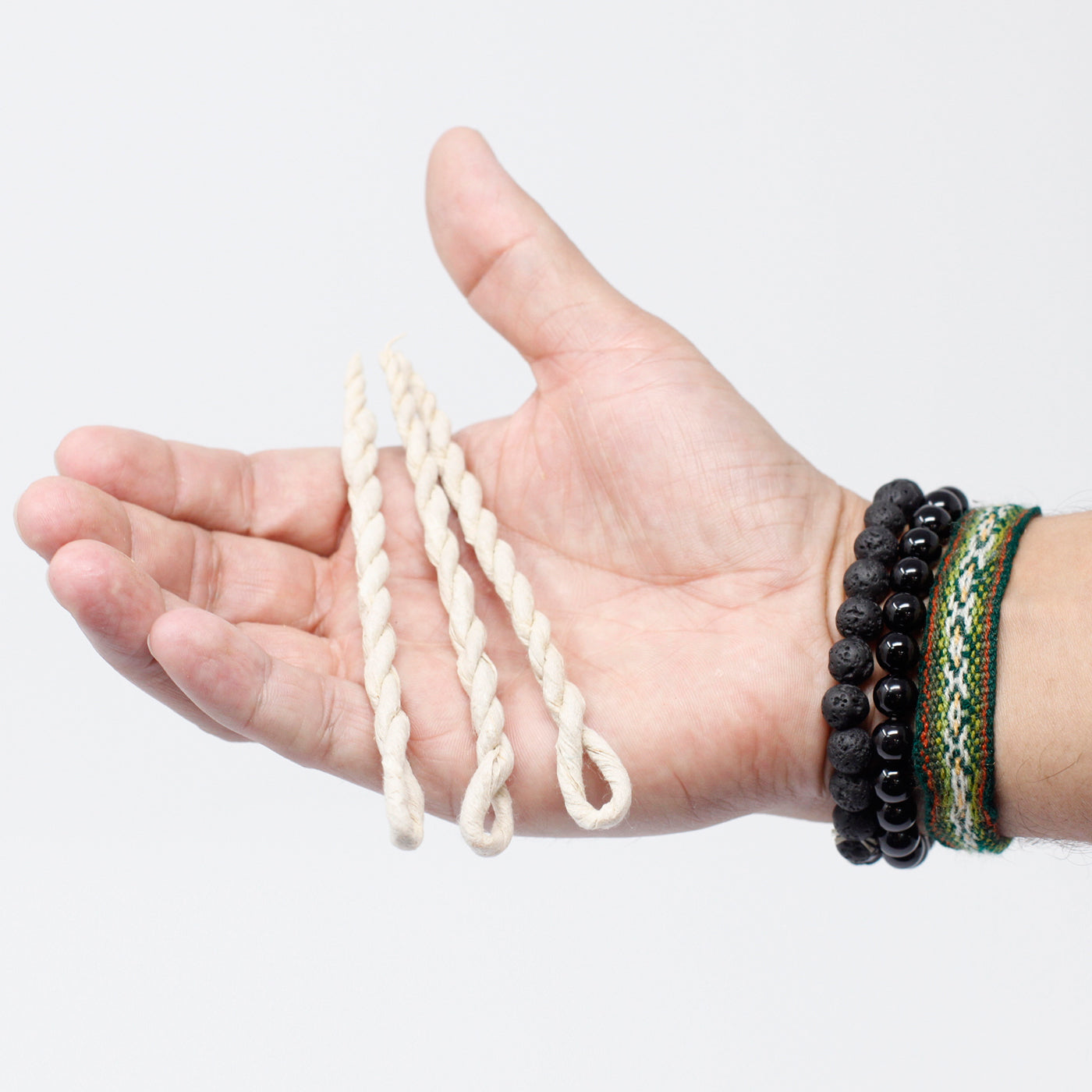 A hand holds three twisted, loop-ended wicks, against a plain background. The person wears black beaded bracelets and a green-patterned wristband.