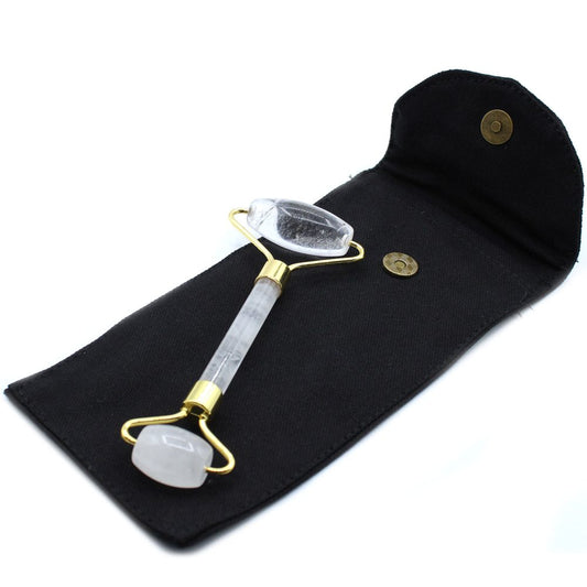 A clear quartz facial roller lays in an open black cloth pouch with a magnetic clasp. The pouch is lying on a white surface.