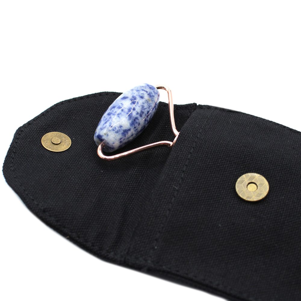 A gemstone facial roller sits inside of an open black carrying case with two brass-colored magnetic clasps. The gemstone is blue and white, and the handle is rose-gold-colored metal.