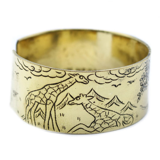 A light gold cuff bracelet features an etched scene of two giraffes in a landscape of mountains, clouds, trees, and birds.