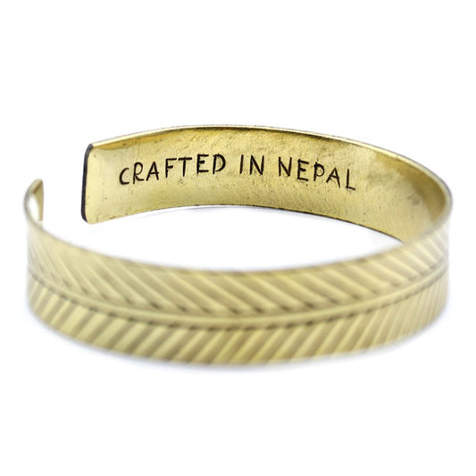 A gold bracelet is displayed on a white surface. The outside shows a feather pattern, while the inside is inscribed with "CRAFTED IN NEPAL."
