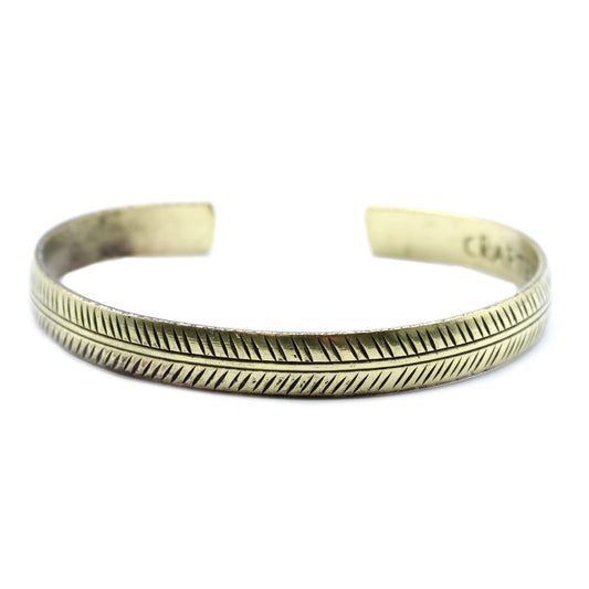 A gold-toned metal cuff bracelet is displayed; the bracelet features a repeating etched pattern and is slightly tarnished. The bracelet is placed on a stark white background.