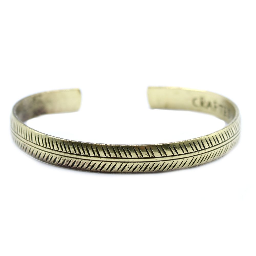 A brass-colored, unfinished metal bracelet with a feathered pattern rests on a plain white background. A blurry engraving is visible inside the bracelet that reads, "CRAFTED".