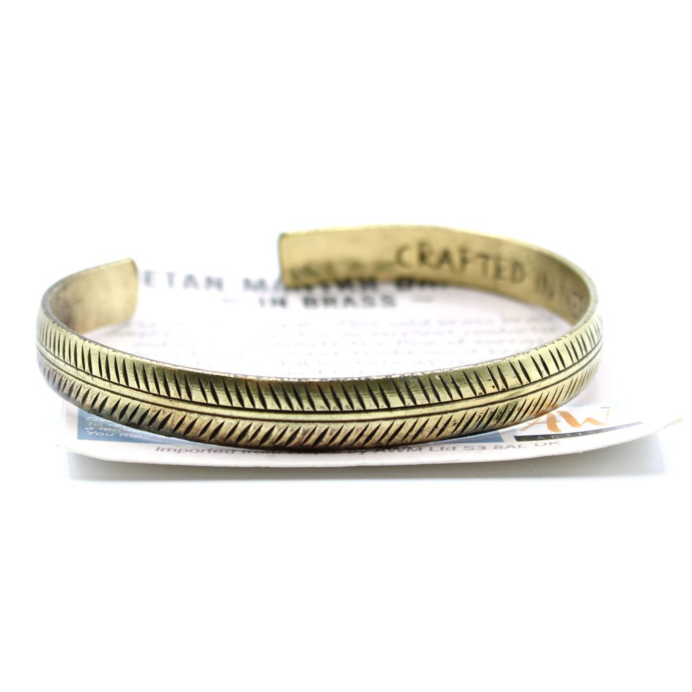 A brass cuff bracelet sits on a white surface, supported by a piece of paper. The bracelet is etched with a feather design and “CRAFTED IN BRASS” is stamped inside. “Imported now by AWM Ltd S3 8AL UK” is printed on the paper.
