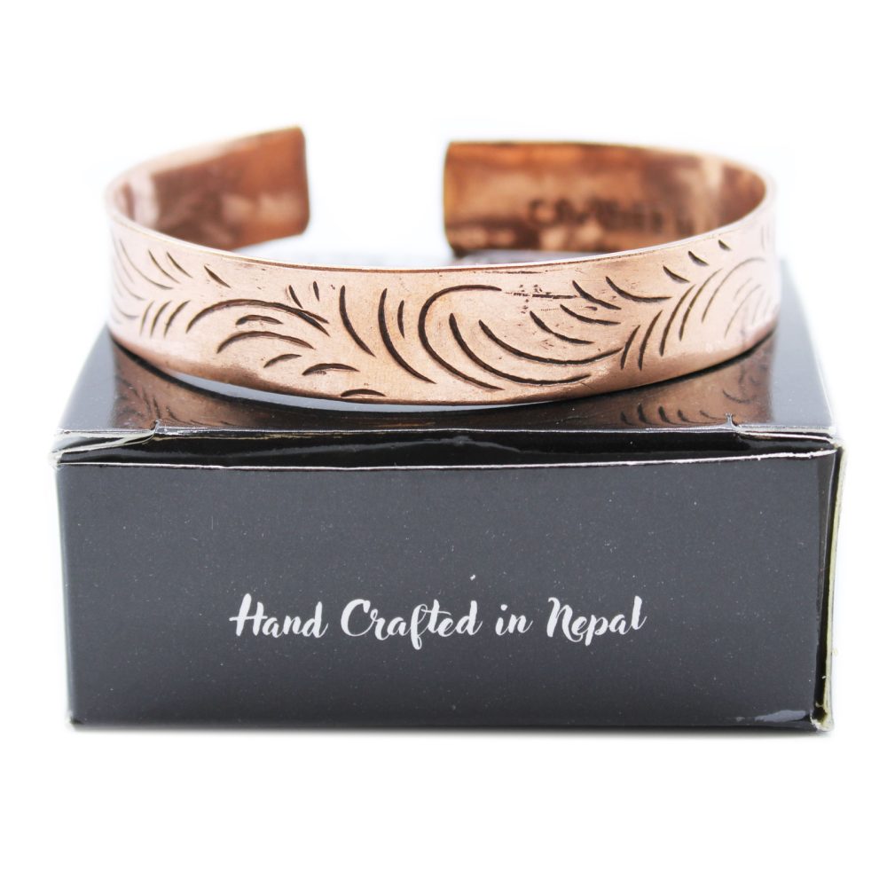A copper cuff bracelet with etched designs rests atop a black box, which displays the text "Hand Crafted in Nepal," against a white background.