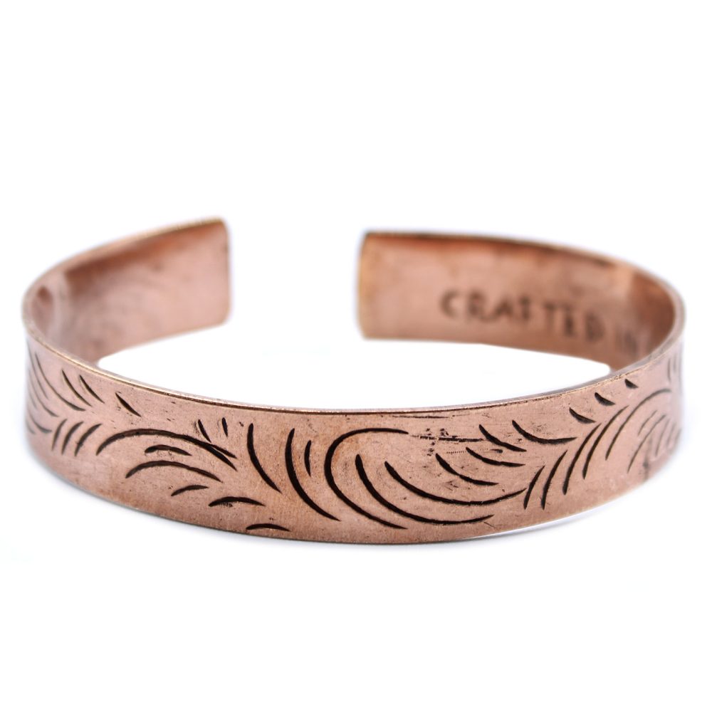 A copper bracelet displays etched floral designs, resting open-ended on a white surface. The word "CRAFTED" is etched on the inside.