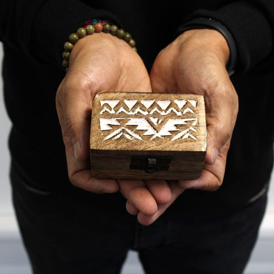 Wooden box with intricate white geometric carvings is held in two hands wearing beaded bracelets. The person is dressed in a black long-sleeved shirt.