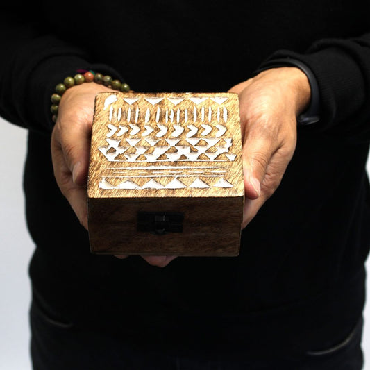 A person holds a wooden box with intricate carved patterns and a clasp, standing against a plain dark background. The person wears a black sweater and a beaded bracelet.