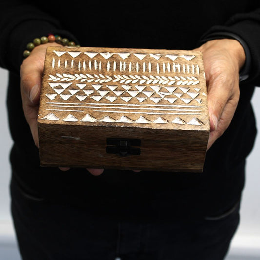A wooden box with white geometric carvings is held in both hands. The person wears a black long-sleeve shirt and a beaded bracelet, against a plain background.