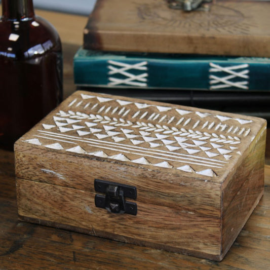 Wooden box with intricate white geometric carvings, closed and secured with a black latch. PosaWhite washed mango wood box with carved Aztec-inspired geometric lid design, hinged construction and dark metal claspitioned on a wooden surface beside a brown bottle and stacked, decorative books.