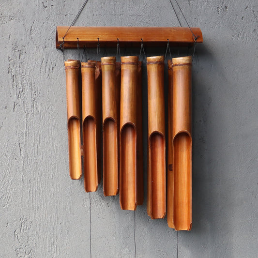 A bamboo wind chime hangs against a gray wall. The chime consists of seven hollow bamboo tubes of varying lengths suspended from a wooden header.