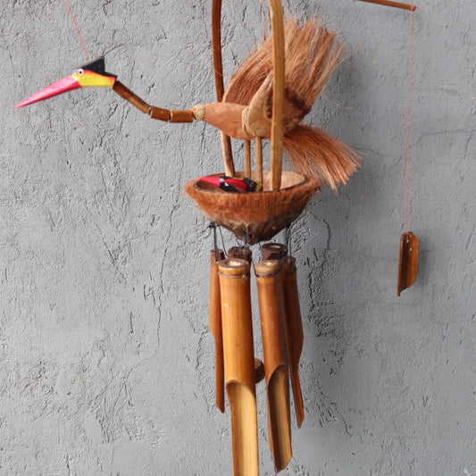 A handcrafted bird-shaped wind chime, made of coconut shell and bamboo, hangs against a textured grey wall. The bird carries a smaller bird in its coconut shell nest.