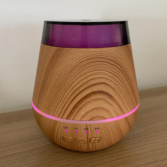 A wood-grain diffuser stands on a wood surface. The top is translucent purple, and a pink light shines between the wood and the buttons labeled "ON 1H 3H 6H."