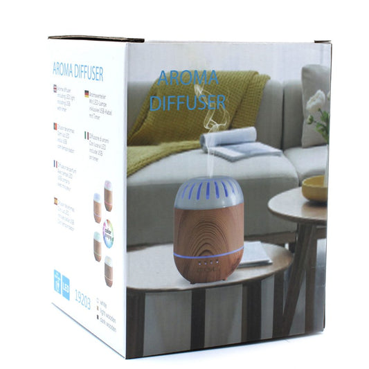 The box for an "AROMA DIFFUSER" is shown. The box features an image of the diffuser emitting steam on a table in a cozy living room setting, along with multilingual text and product specifications, "including LED light, including USB, with timer", "color changing," and color swatches.