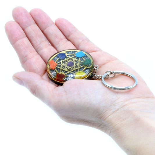 A circular keychain with inlaid gemstones sits on an open palm. A silver ring is attached, and the background is a plain white.