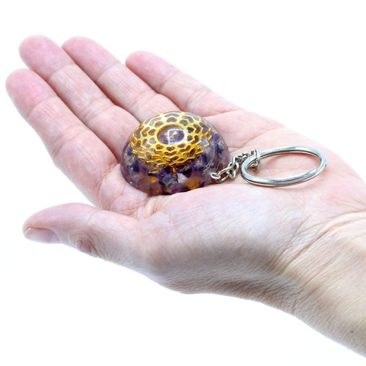 A purple crystal keychain rests in an open palm, its metallic golden top catching the light; a ring is connected to the keychain. The background is a bright white.