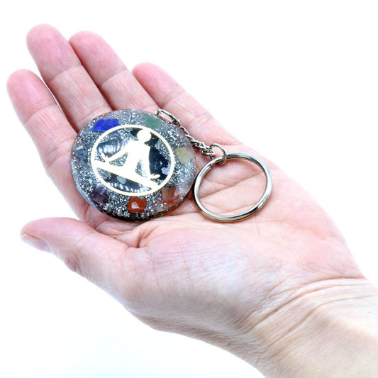A round, silver keychain with a human figure is held in a palm, the background being a bright white. The keychain has several colorful stones around the edge.