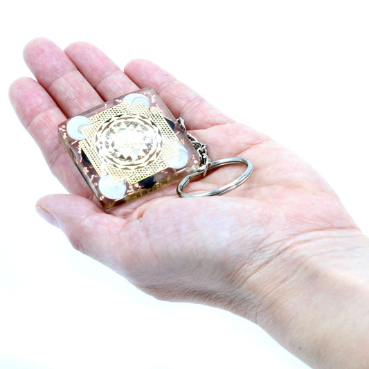 A clear, square keychain with gold designs and a metal ring rests in the palm of an outstretched hand against a plain white background.