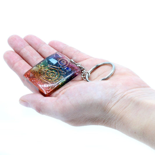 A resin keychain with embedded stones, a coil, and a metal ring rests on a person’s outstretched palm against a bright white background.