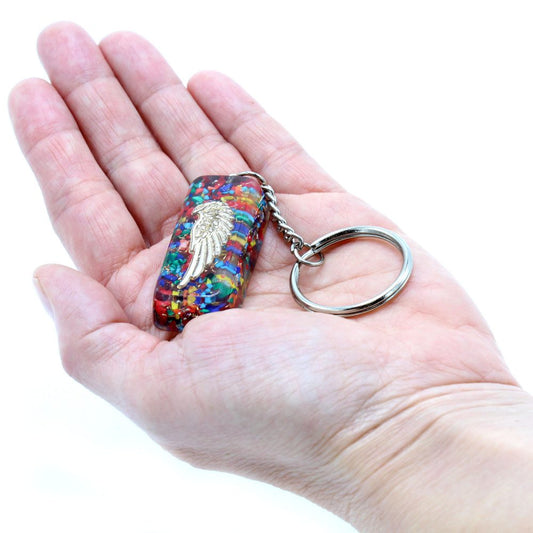 A colorful, rectangular keychain with a gold-colored winged emblem rests in an open palm, connected to a silver key ring, all set against a bright white backdrop.