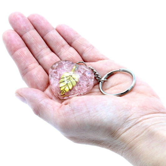A pink, heart-shaped keychain with a gold leaf design rests in the palm of a person's open hand against a plain white background.