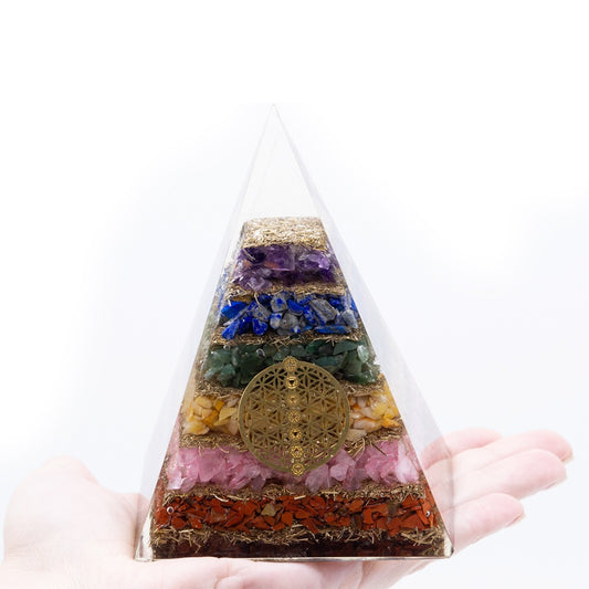 A pyramid-shaped orgonite is held in a palm, showing layers of colored crystals separated by metallic fillings, and accented with a gold-colored flower of life symbol, against a plain white background.