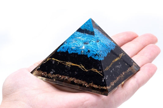 A resin pyramid, layered with gold flakes, black material, and blue stones, sits on an open palm. The background is a solid white.