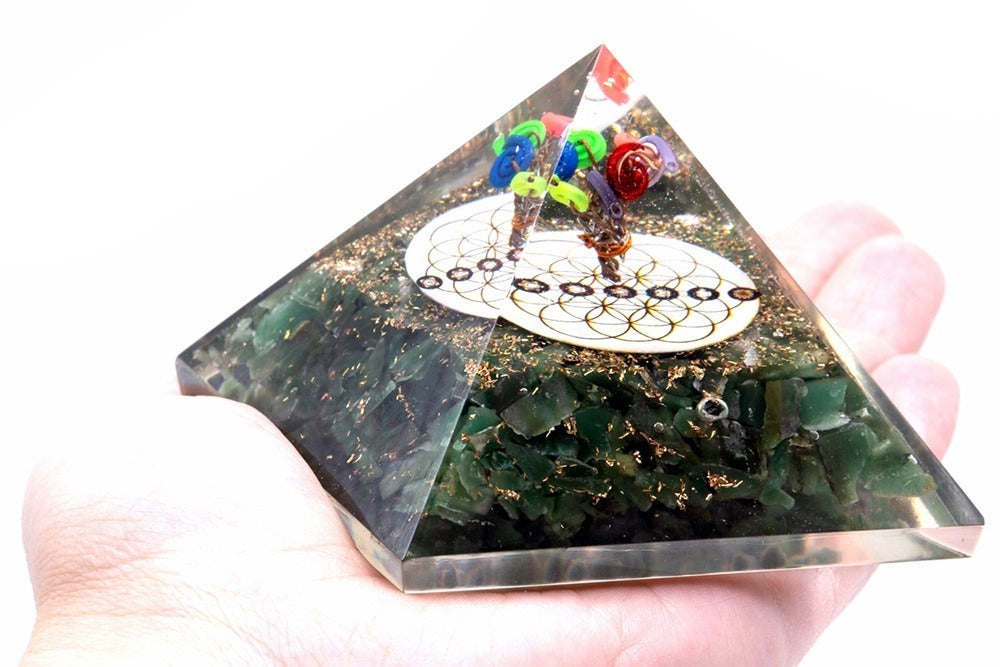 A pyramid-shaped orgonite with embedded green stones, gold flakes, a tree with colorful "leaves", and "Flower of Life" symbol sits in the palm of a hand against a white background.