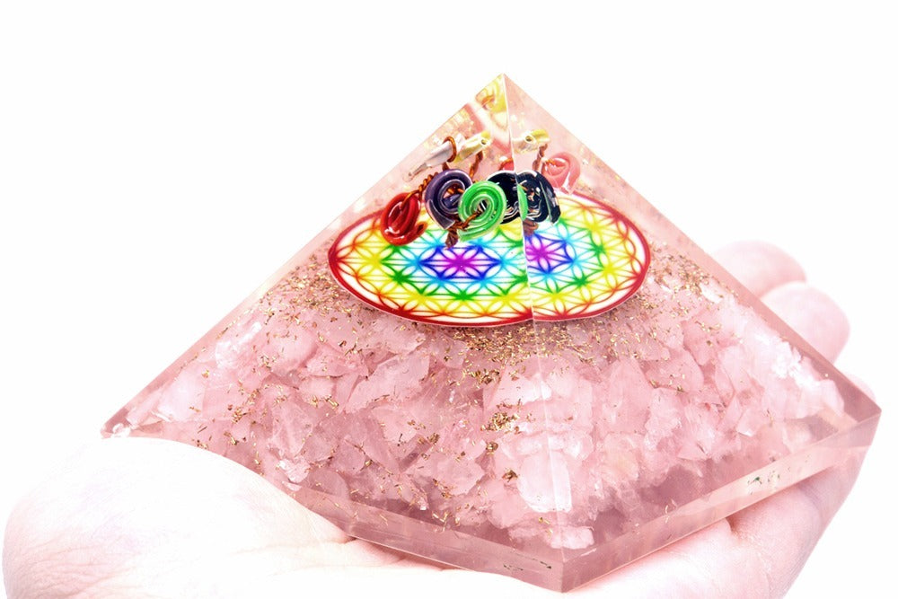 A pyramid made of pink crystals and gold flakes sits in a hand. The top has a colorful "Flower of Life" symbol and metal coils. The background is white.