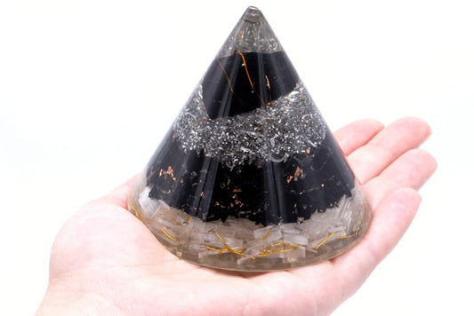 A layered orgone pyramid rests on a palm. The pyramid contains layers of silver-colored metal shavings, black stones, and clear crystals, encased in a translucent resin. The background is white.