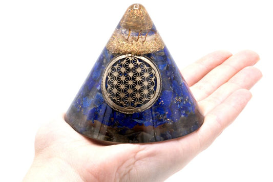 A blue, cone-shaped orgone pyramid with a metallic "flower of life" symbol is held in an open hand, displayed against a white backdrop.