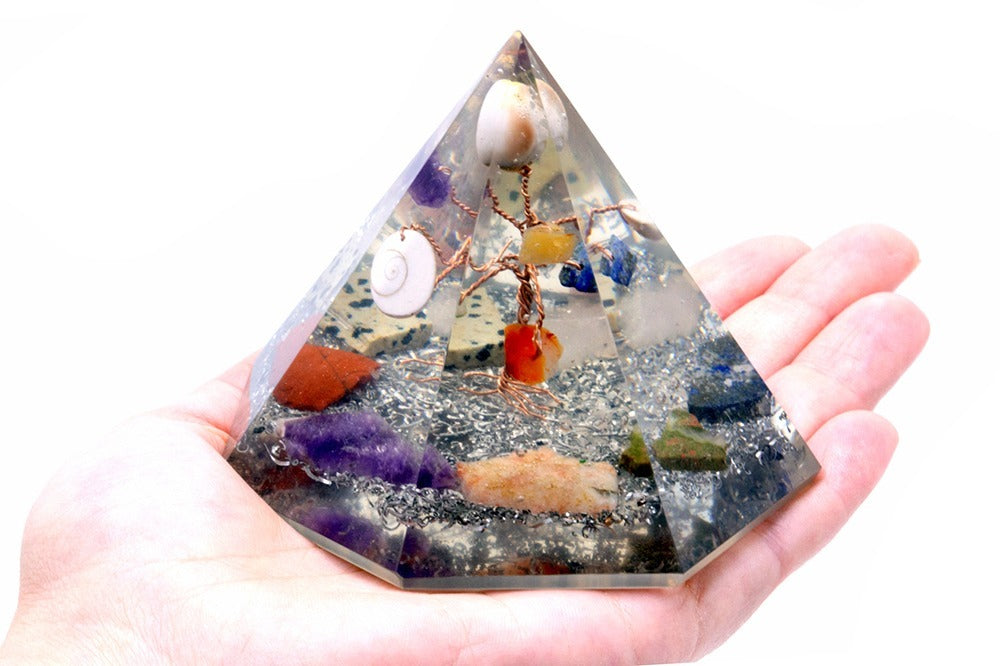 A clear pyramid, filled with crystals, metal shavings, and a wire tree with shells, rests in an open human palm. The background is white.