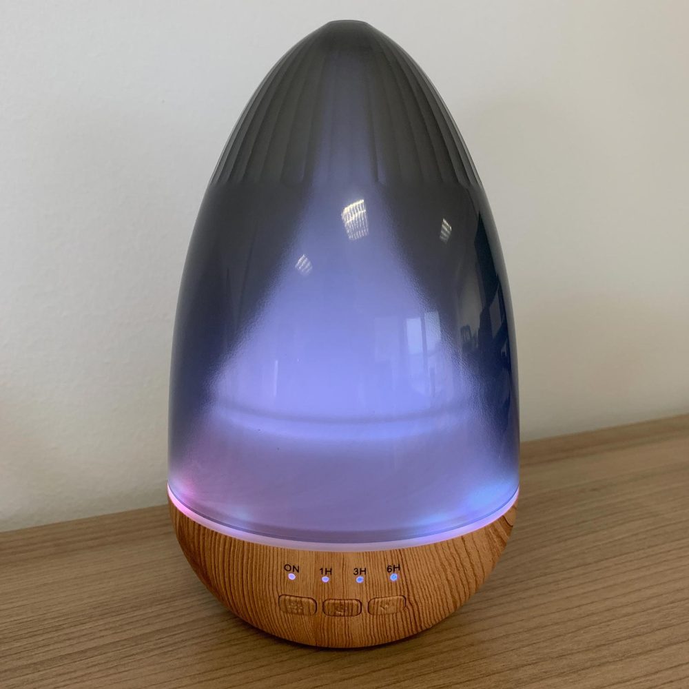 A gray and wooden aromatherapy diffuser sits on a wooden surface, emitting a lavender light from its center. Buttons labeled "ON," "1H," "3H," and "6H" are visible on the wooden base.