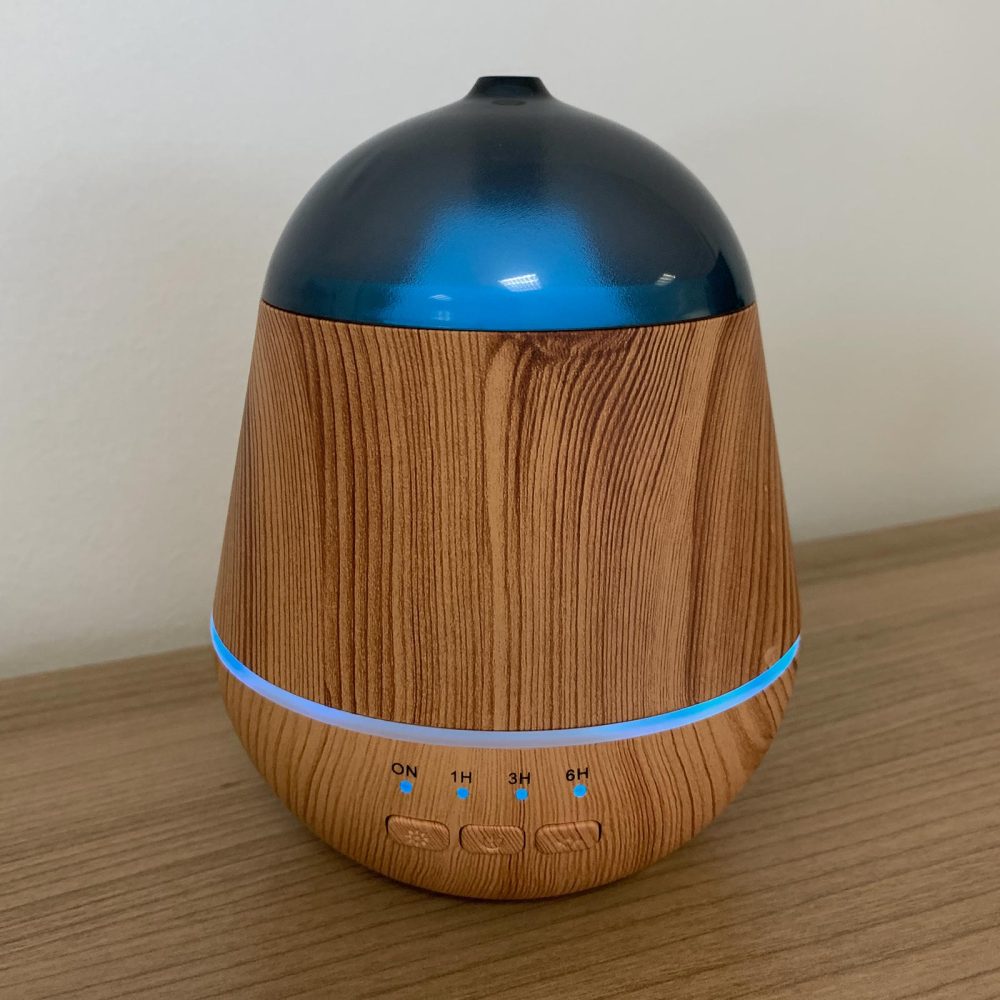 A wood-look diffuser stands on a wooden surface. It glows with a blue light and has buttons labeled "ON," "1H," "3H," and "6H."