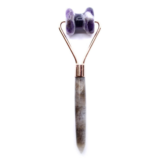 A face roller with a two-pronged rose gold frame holds purple stone rollers above a grey stone handle, set against a plain white background.