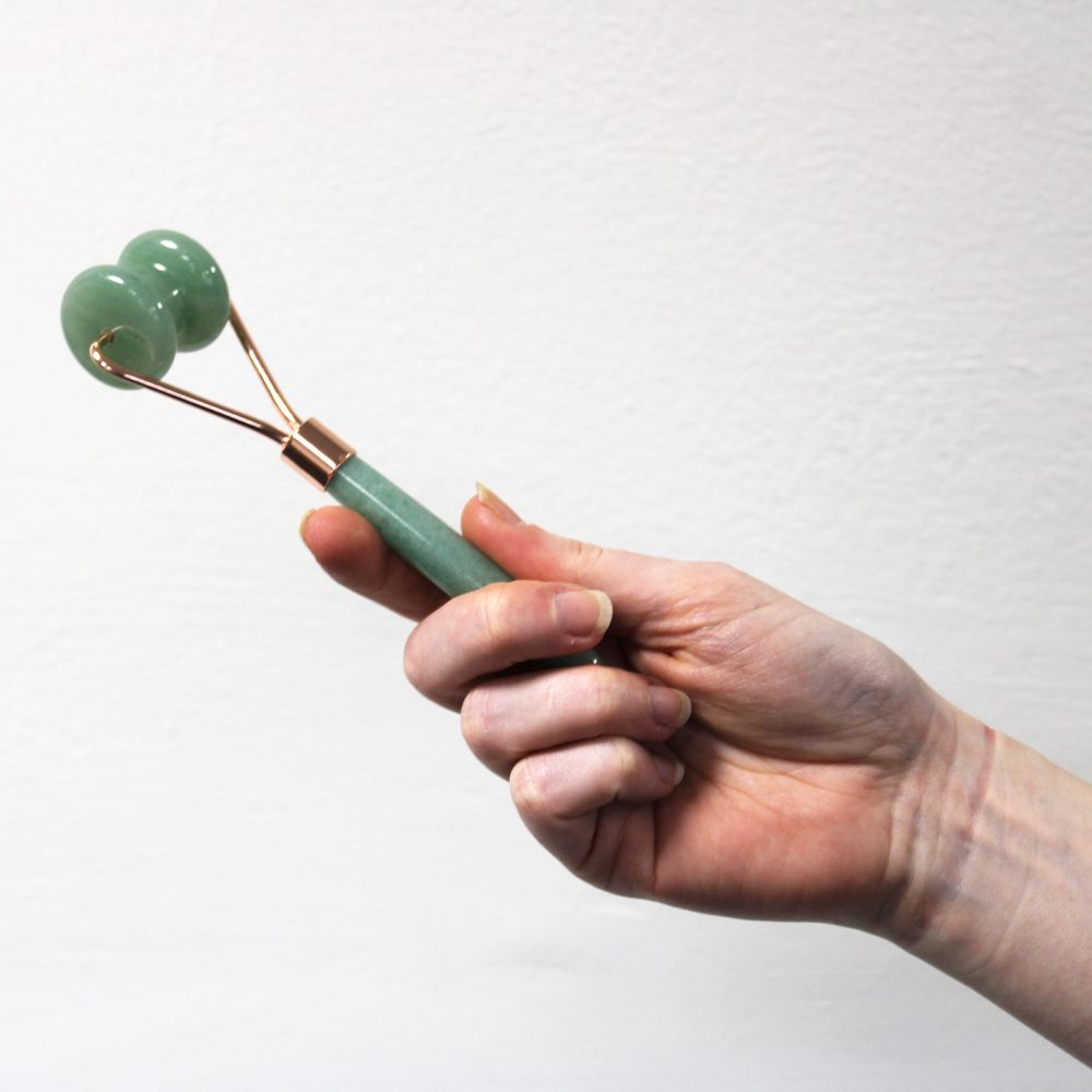 A fair-skinned hand holds a double-roller jade face massager with copper supports. The background is a plain, textured white.