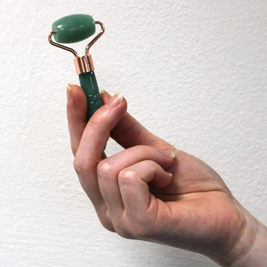 A hand holds a green gemstone facial roller with a copper-colored metal frame. The background is a textured white wall.