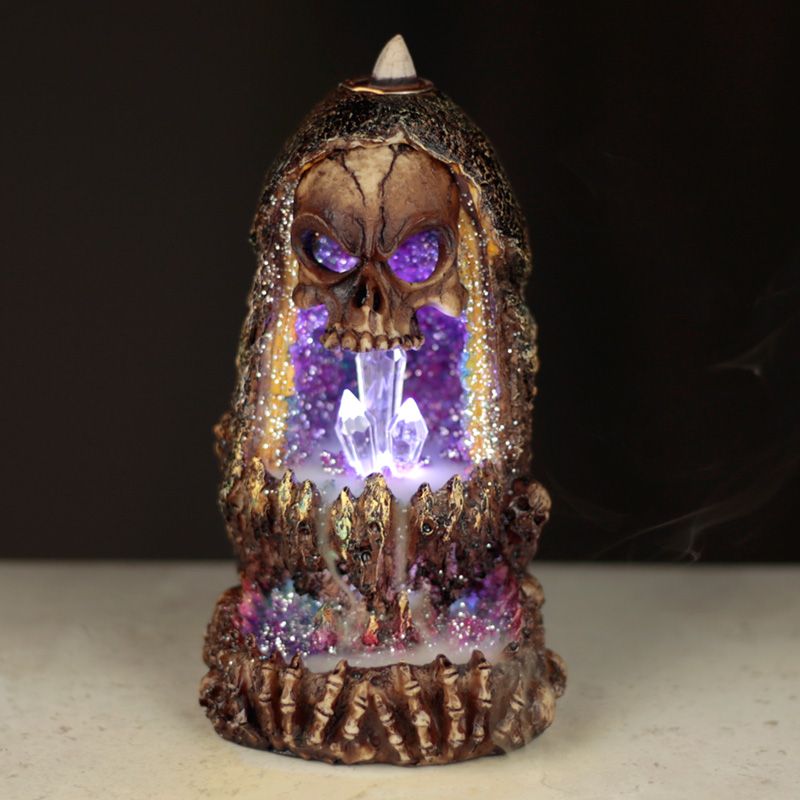 A skull-adorned incense burner releases smoke, illuminating glowing crystals inside. The background is dark, highlighting the mystical atmosphere created by the colors and eerie design.