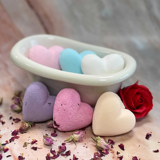 Heart-shaped bath bombs rest in a small, white container, surrounded by scattered dried rose petals and a single red rose on a marble surface.