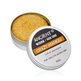 A round, solid shampoo bar sits in an opened, circular metal tin. Beside it is the tin’s lid that reads: "ANCIENT WISDOM/HAIR CARE CRAZY MANGO Solid shampoo FOR HEALTHY, SHINY HAIR 60G."