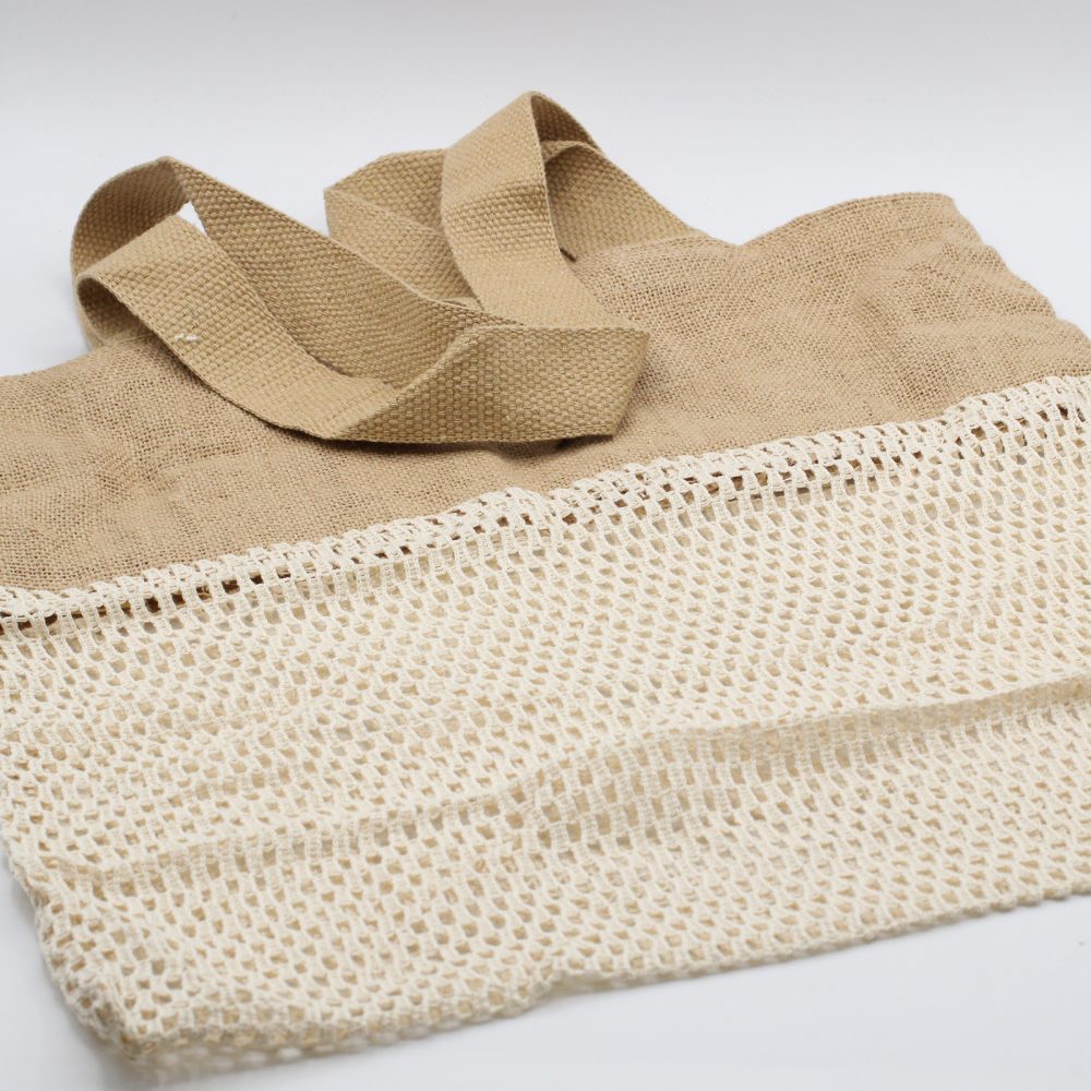 A tan tote bag sits on a white surface. The top portion of the bag is woven fabric, and the bottom portion is open weave. It has sturdy matching handles.