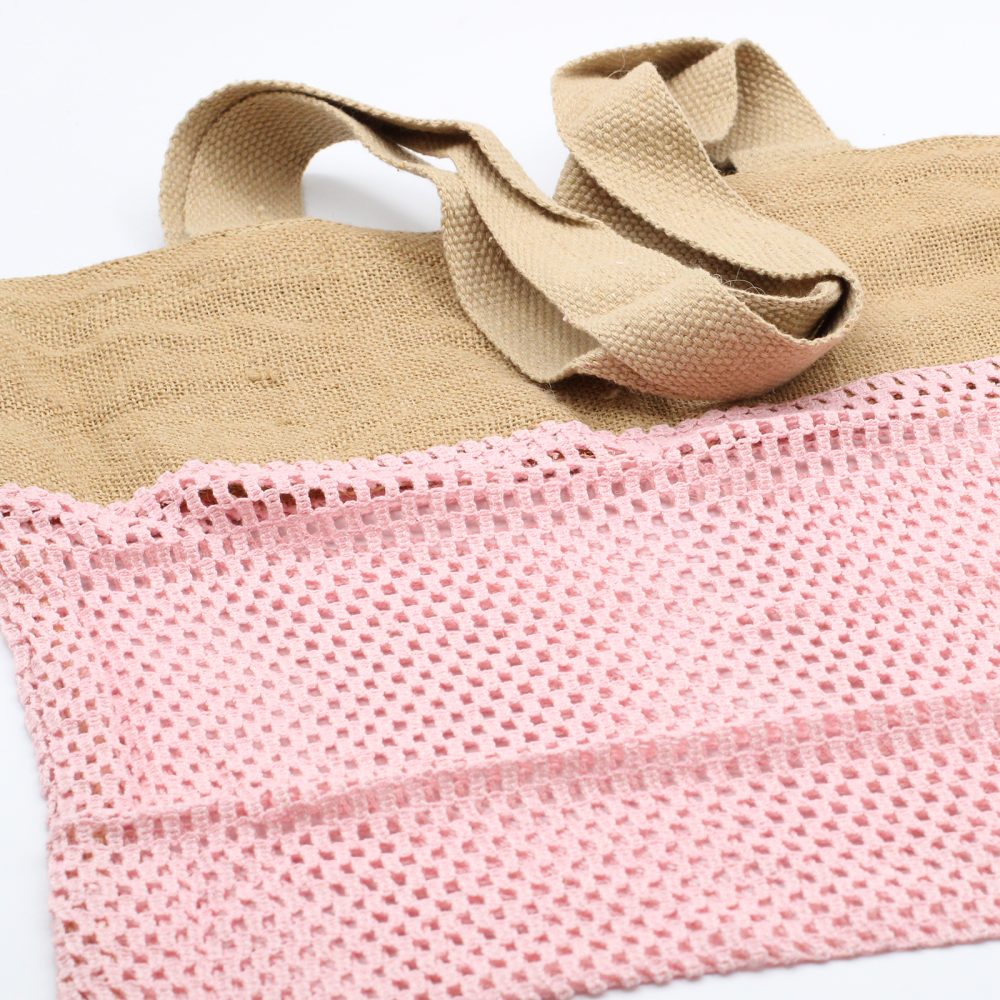 A beige tote bag with handles rests on a white surface. The bottom of the bag features pink netting.