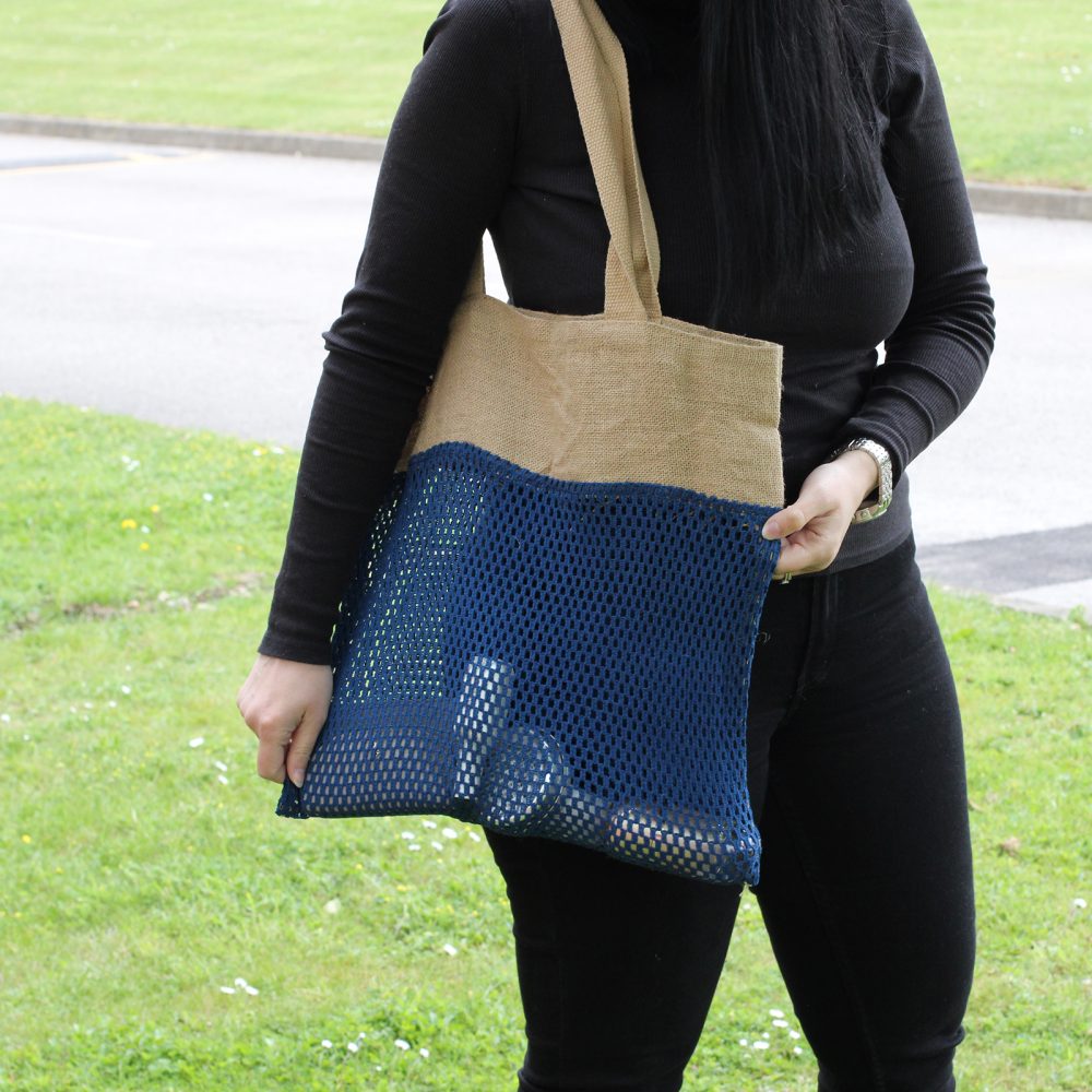 A woman holds a jute and blue mesh tote bag. She is on a grassy lawn with a roadway behind her.
