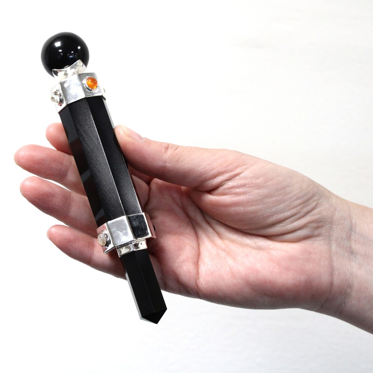 A hand holds a decorative black wand with a polished orb on top. It has silver fittings with small, embedded, colored gems. The backdrop is white.