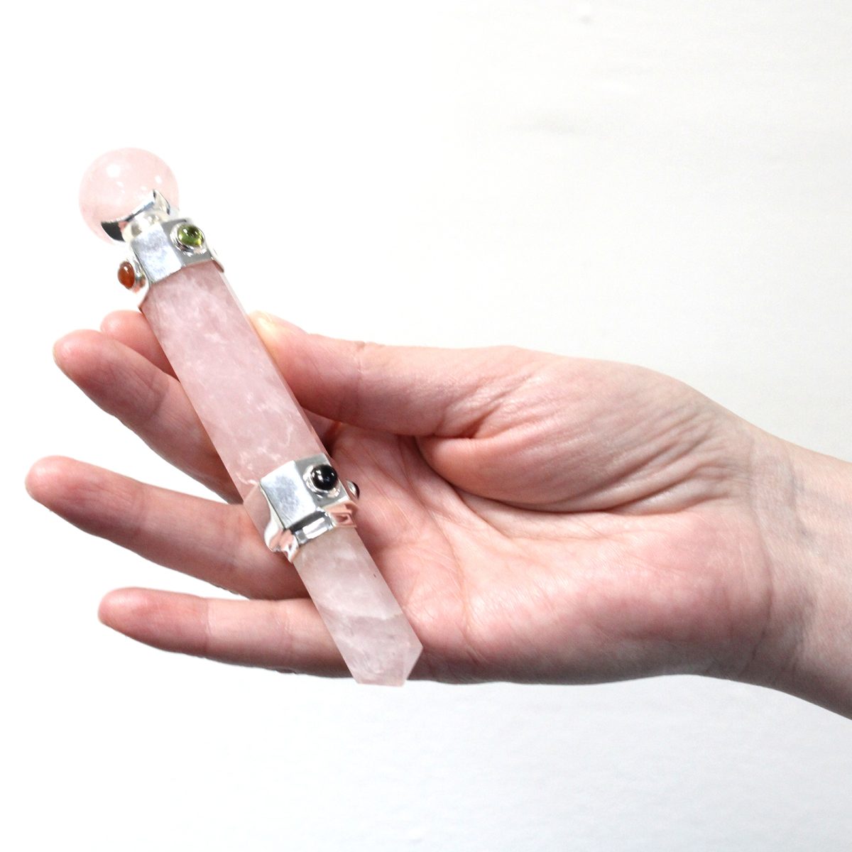A rose quartz wand, adorned with decorative metal and other stones, rests in the palm of a fair-skinned hand, against a plain white background.