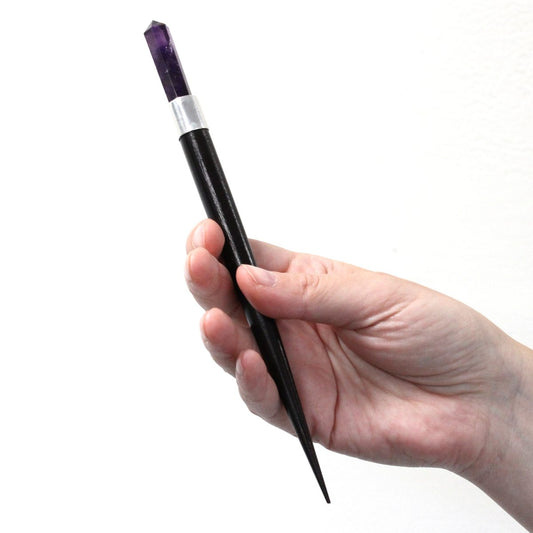 A hand holds a long, dark wand with a pointed end and an amethyst crystal at the opposite end, set against a plain white background.